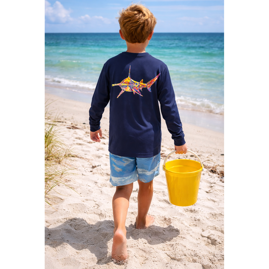 Child walking on a beach with a yellow bucket, wearing a navy long-sleeve sun shirt with a colorful hammerhead shark design.