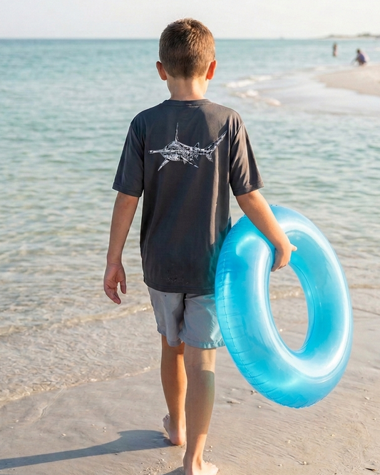 Child walking on a beach holding a blue inner tube with a gray short sleeve Hammerhead sun shirt with the ocean and sky in the background
