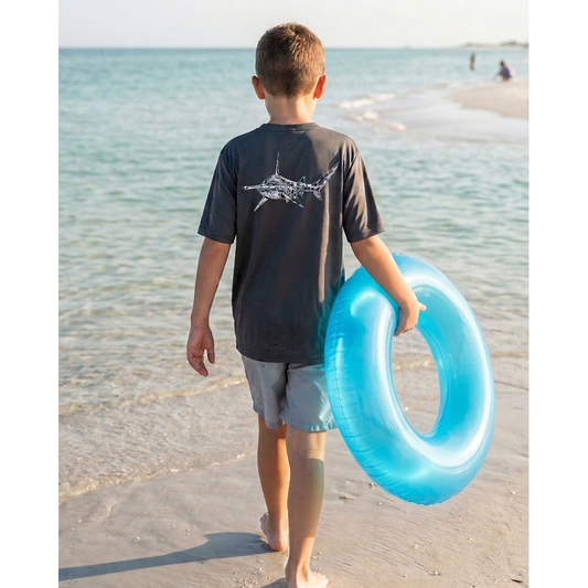 Child walking on a beach holding a blue inner tube with a gray short sleeve Hammerhead sun shirt with the ocean and sky in the background