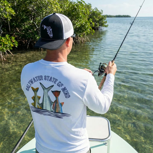 Fisherman wearing white long sleeve performance UV shirt with Inshore Slam fish tails (snook, tarpon, redfish) and text "Saltwater State of Mind", fishing on a boat in the mangroves.