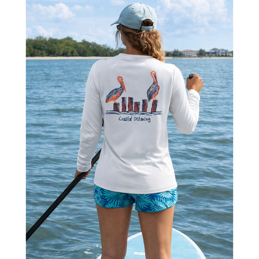 Lady paddleboarding on a lake wearing a white long-sleeve sun shirt with pelican design and the text "Coastal Distancing".