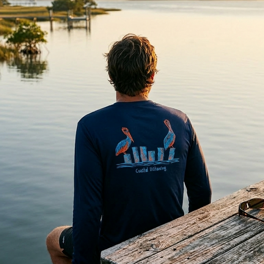 Model wearing a navy blue long sleeve UPF 50 sun shirt with 2 pelicans on stumps on the back and the text "Coastal Distancing"