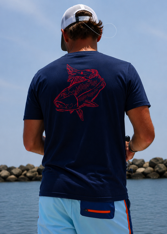 Back view of a navy Red Drum cotton t-shirt featuring a large redfish graphic, worn by an angler fishing from a boat along a rocky shoreline.