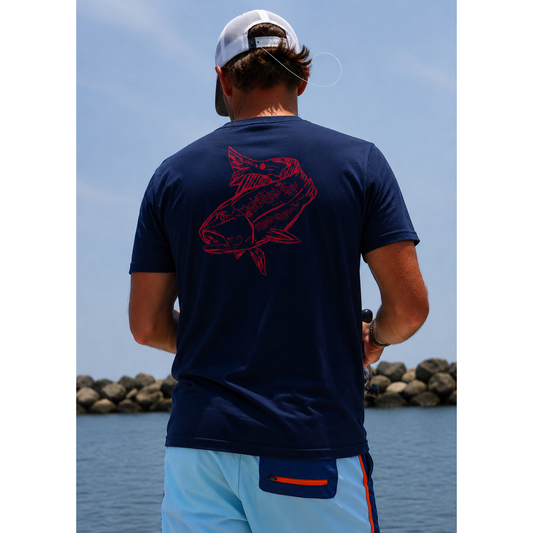 Back view of a navy Red Drum cotton t-shirt featuring a large redfish graphic, worn by an angler fishing from a boat along a rocky shoreline.