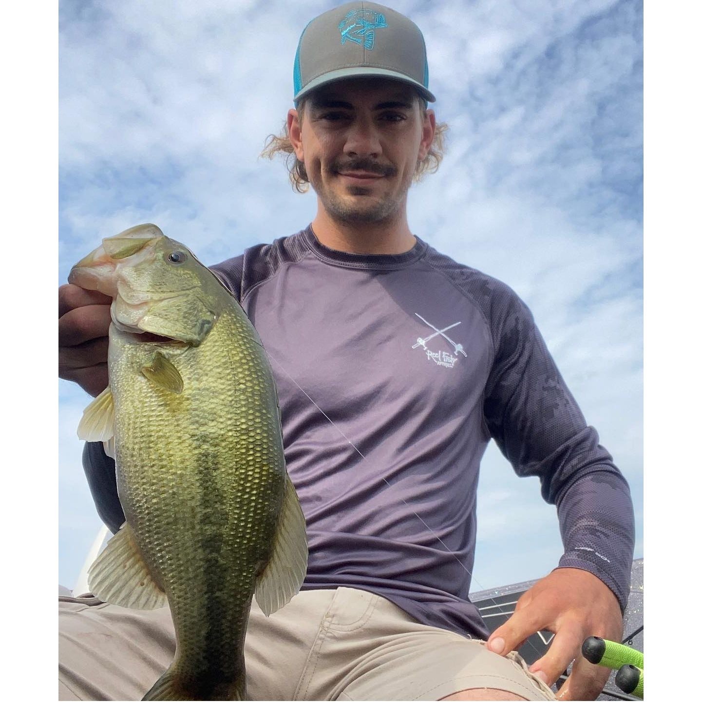 Fisherman holding a bass while wearing a "Reel Hawg" graphite digital camo long sleeve performance fishing shirt with 50+ UV protection