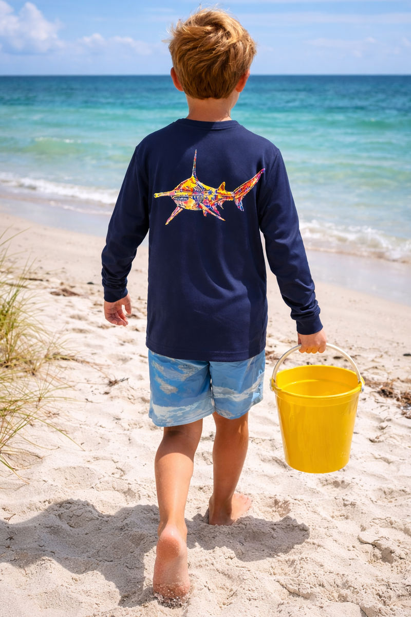 Child walking on a beach with a yellow bucket, wearing a navy long-sleeve sun shirt with a colorful hammerhead shark design.