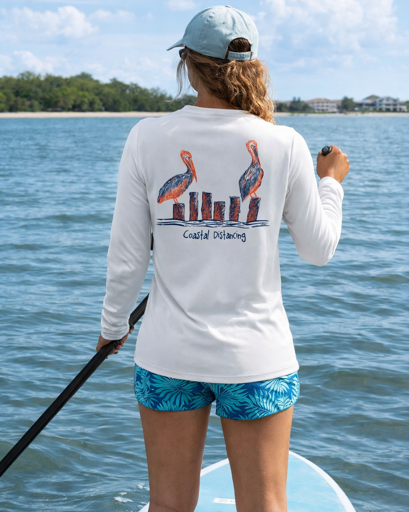 Lady paddleboarding on a lake wearing a white long-sleeve sun shirt with pelican design and the text "Coastal Distancing".