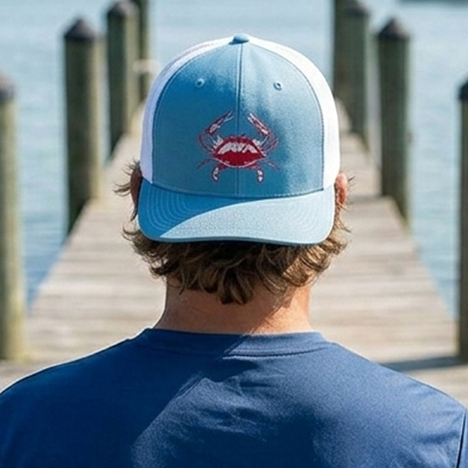 Guy wearing a light blue trucker hat with a blue crab embroidery on a dock