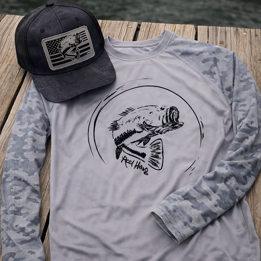 Bass fishing trucker hat in Kryptek black Richardson 112 with laser-engraved USA flag bass leather patch resting on a gray UPF 50+ performance long sleeve fishing shirt with camo sleeves, laid flat on a wooden dock.