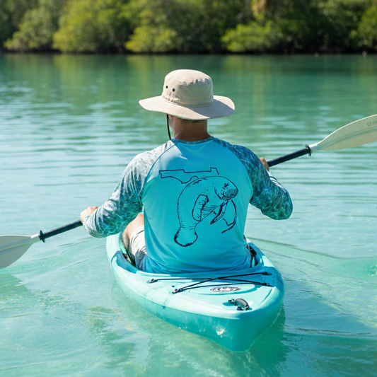 Back view of a man kayaking in calm water wearing an aqua blue performance fishing shirt with water camo long sleeves and a manatee with Florida state graphic on the back, paired with a wide-brim booney sun hat.