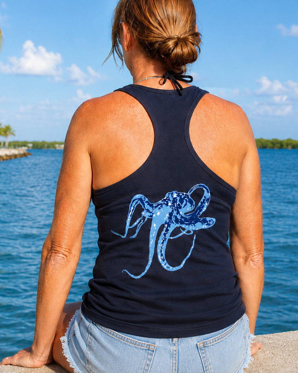 Model wearing a navy cotton racerback tank top with a blue octopus design on the back while sitting on a wall over the water