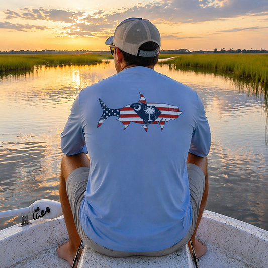Man sitting on a boat wearing a light blue UPF 50 performance shirt with a Tarpon fish and the State of South Carolina Flag design, looking at a sunset over water.