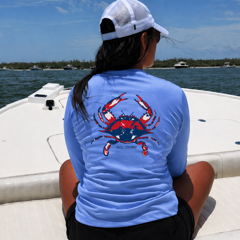 A woman sitting on the bow of a boat, viewed from behind, wearing a light blue long-sleeve performance shirt with a red and blue crab graphic on the back. Text "Reel Crabby" is printed below the graphic.