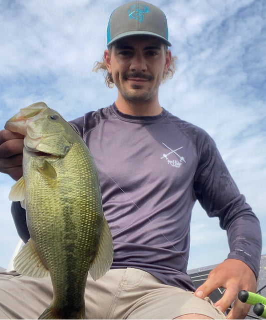 Fisherman holding a bass while wearing a "Reel Hawg" graphite digital camo long sleeve performance fishing shirt with 50+ UV protection
